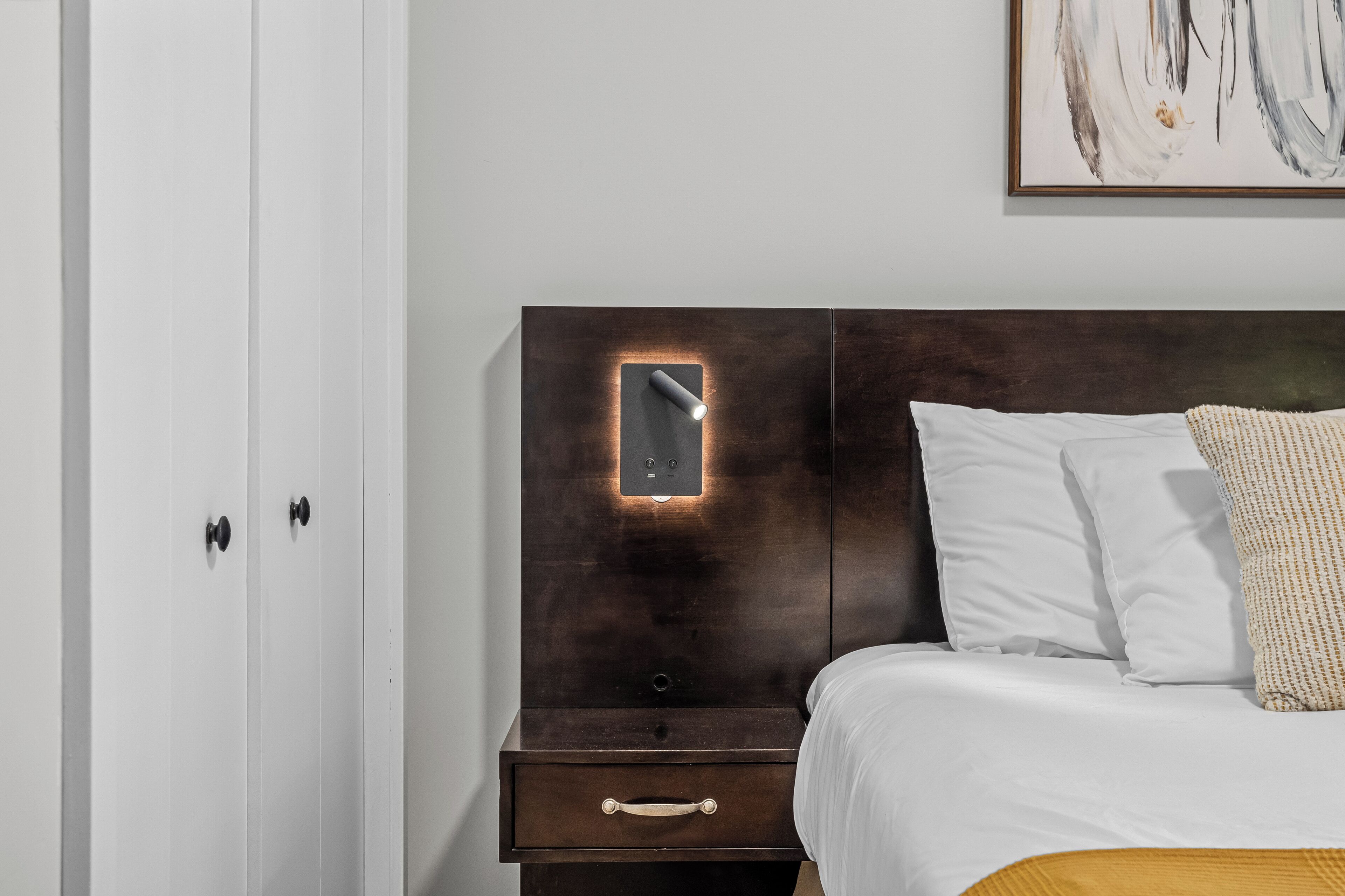A bedroom with a dark wooden headboard, a bedside table with a drawer, and a white bed with a textured pillow.