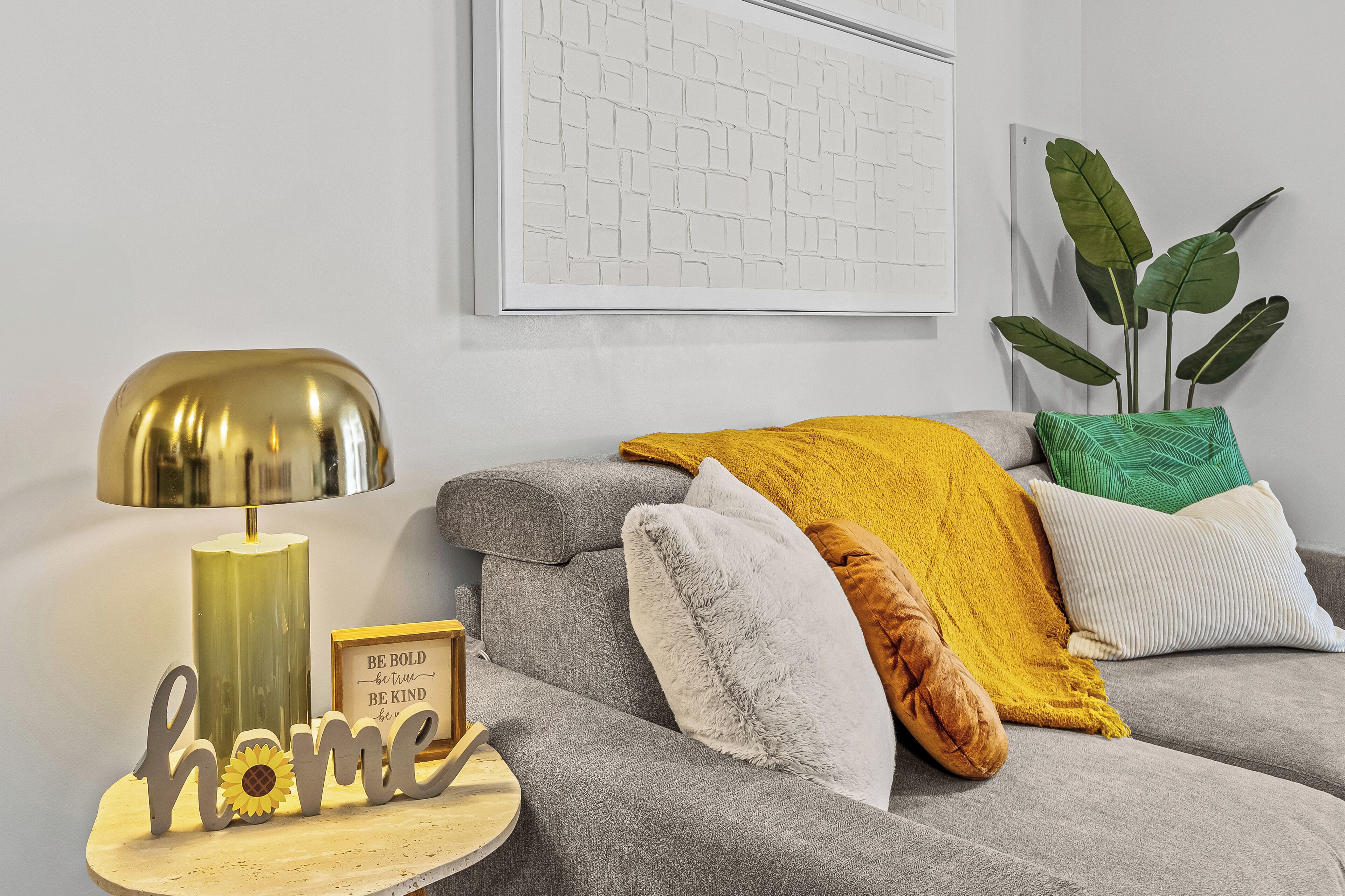 Ein modernes Wohnzimmer mit einem grauen Sofa, einem runden Beistelltisch, einer goldfarbenen Lampe und dekorativen Kissen.
