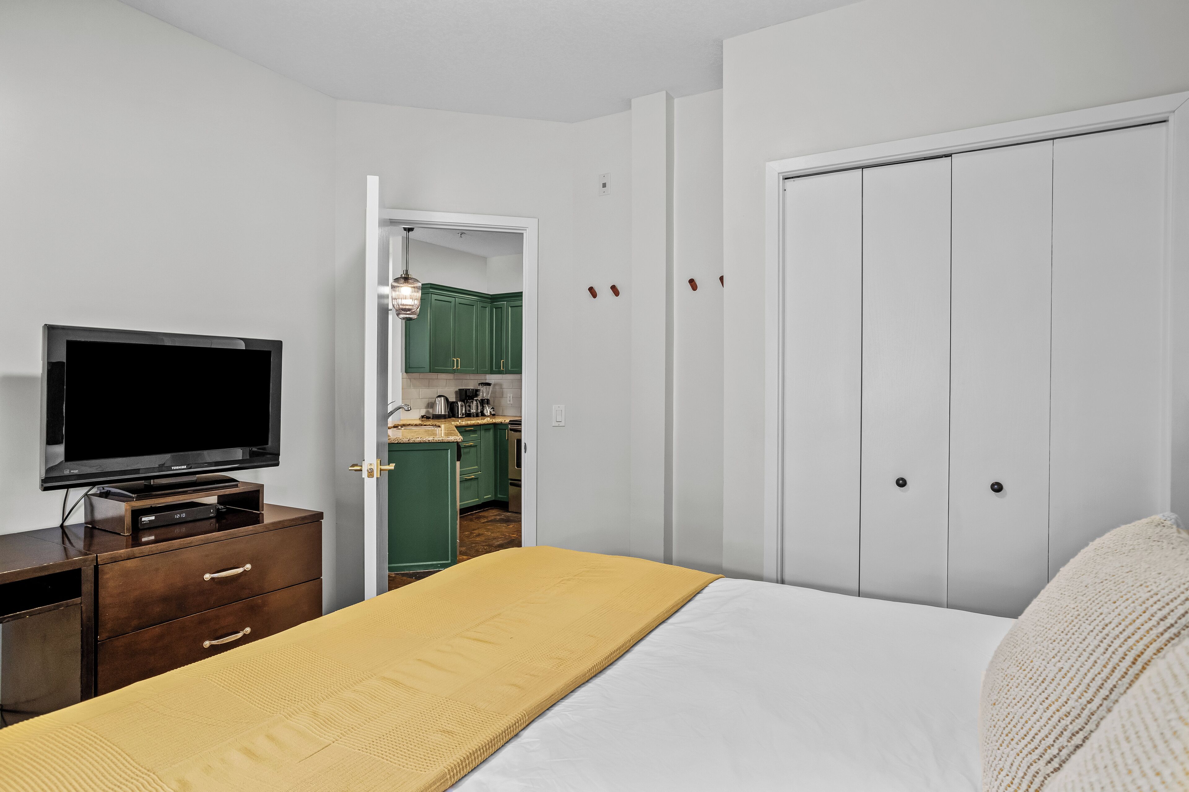Une chambre à coucher avec un lit, un téléviseur posé sur une commode, et une cuisine visible à travers une porte ouverte.