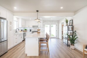 A modern kitchen with white cabinets, a central island, and a dining area with wooden chairs. - Horne's Haven, A Quiet Cozy Home (Oceanside)