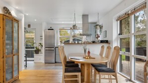 Et moderne køkken med en central ø, et spiseområde med træstole og et rundt bord, samt store vinduer med udsigt til det fri.