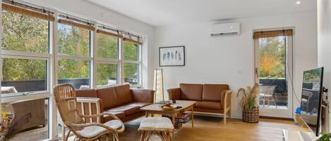 A spacious living room with a brown leather sofa, wicker chairs, and a wooden coffee table. Large windows offer views of trees and a deck outside.
