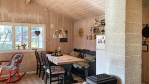 Ein gemütliches Holzinnere mit Esstisch, Stühlen, Schaukelstuhl, Kamin und einer Uhr an der Wand.