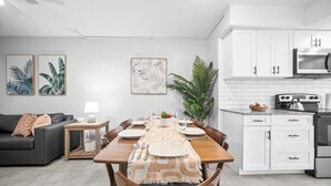A modern kitchen with white cabinets, a microwave, and a stove. There is a dining area with a wooden table set for four, a sofa, and a small side table with a lamp. - Holiday Park by Lowkl (Fort Lauderdale)