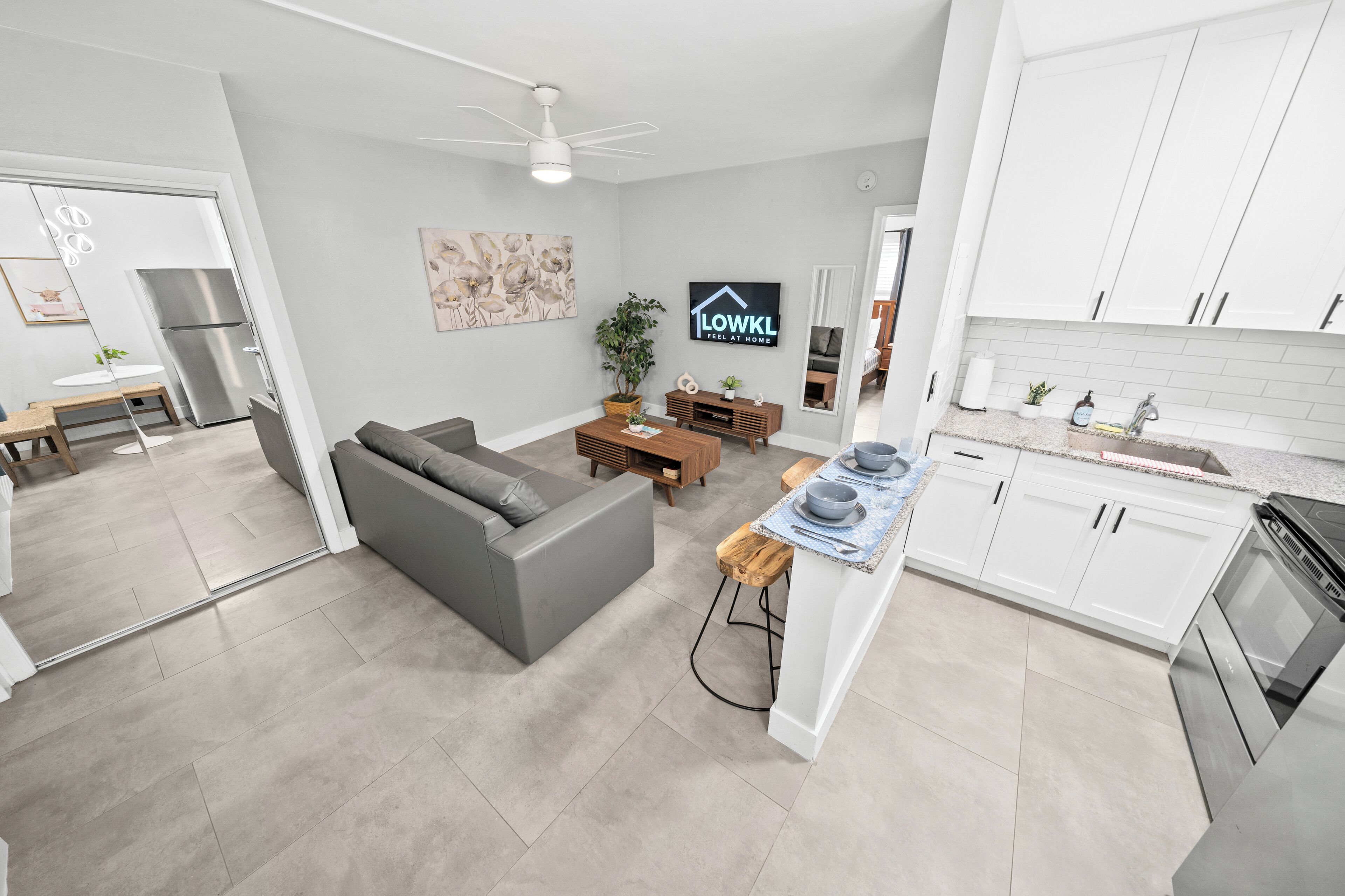 A modern apartment with a grey sofa, wooden coffee table, and a kitchen with white cabinets and a stainless steel refrigerator.