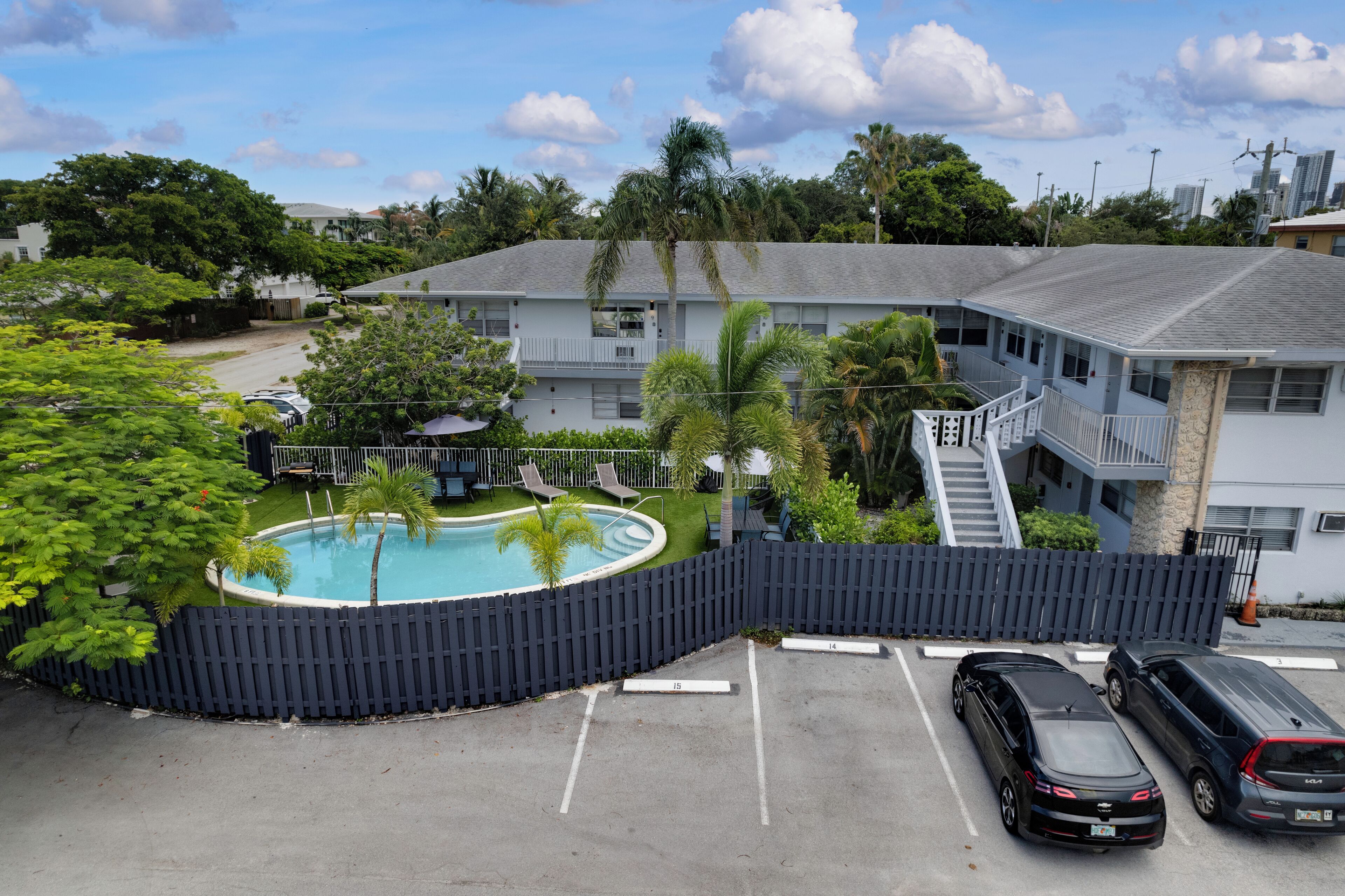 A pool area with a fence, a parking lot with two cars, and a building with balconies and a staircase.