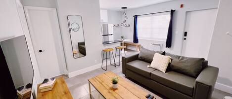 A modern living room with a sofa, coffee table, and a TV. There is a kitchen area with a fridge and a dining table.