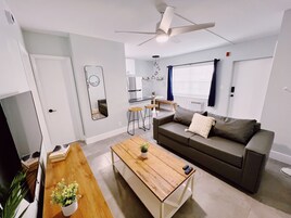 Una sala de estar moderna con un sofá, una mesa de centro y un televisor. Hay una zona de cocina con un refrigerador y una mesa de comedor.
