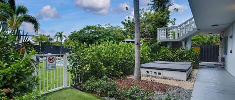 A modern outdoor area with a white gate, lush greenery, a palm tree, and a small building with a flat roof.