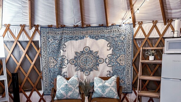 A cozy interior with a bed, ottomans, a patterned tapestry, and a wooden shelf with a plant.
