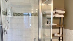 A modern bathroom with a glass-enclosed shower, white tiled walls, a toilet, and a shelf with towels.