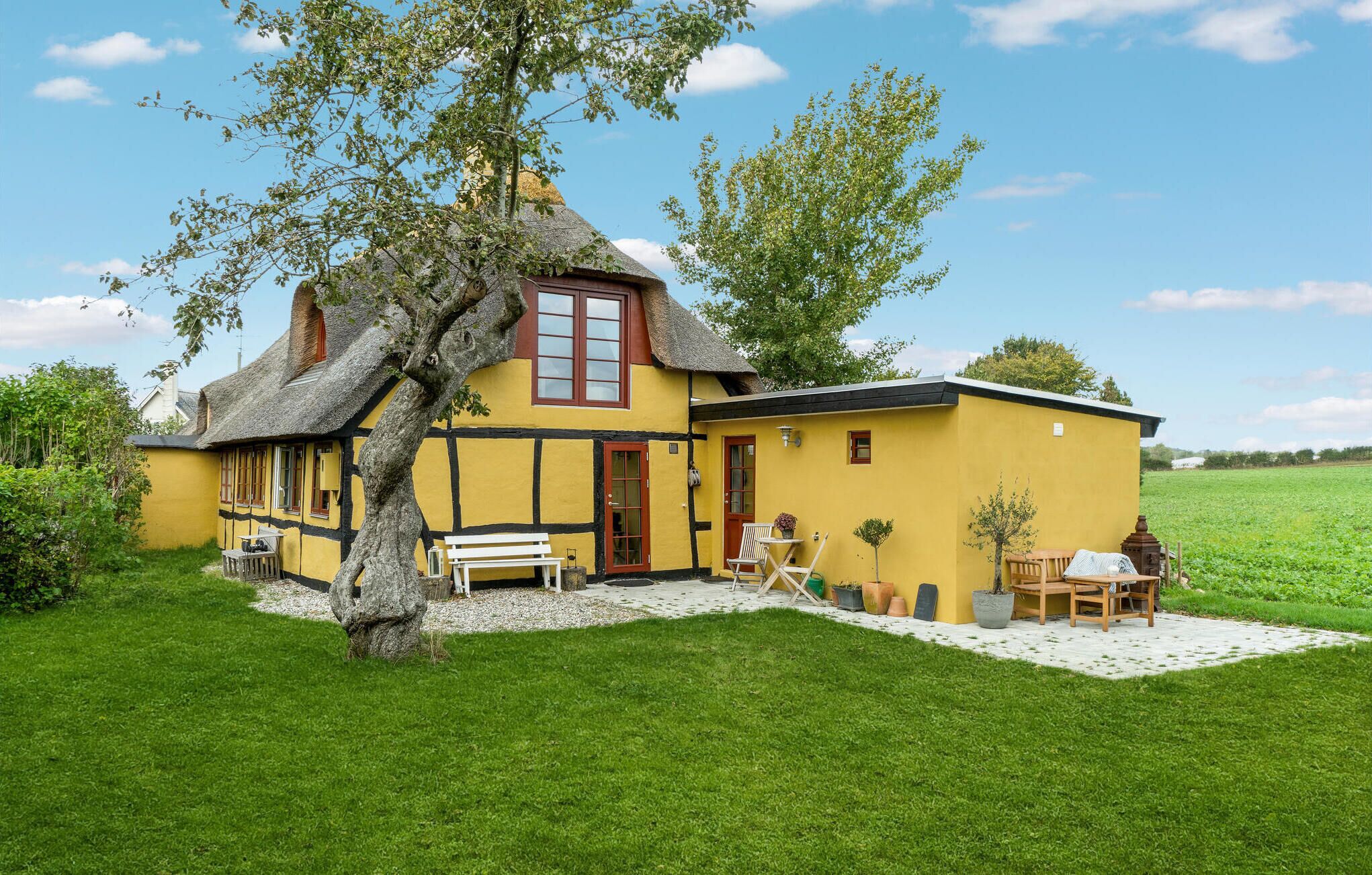 Una casa tradicional amarilla con techo de paja, un gran árbol en el frente y un pequeño jardín con muebles de exterior.