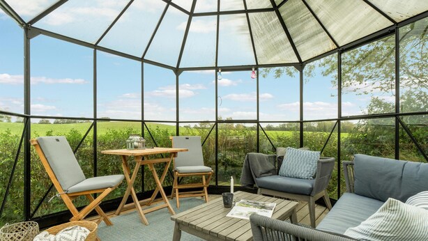A glass-enclosed room with a wooden table, two chairs, a sofa, and a view of a green field. - Lovely home in Rudkøbing with WiFi (Rudkøbing)