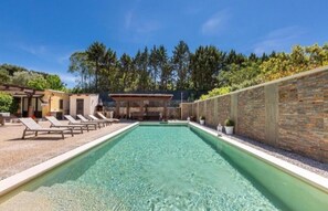 Ein rechteckiger Pool mit klarem Wasser, umgeben von einem mit Kieselsteinen ausgelegten Deck. Entlang des Poolrandes stehen Liegestühle, und im Hintergrund befindet sich ein überdachter Sitzbereich mit Tisch und Stühlen.