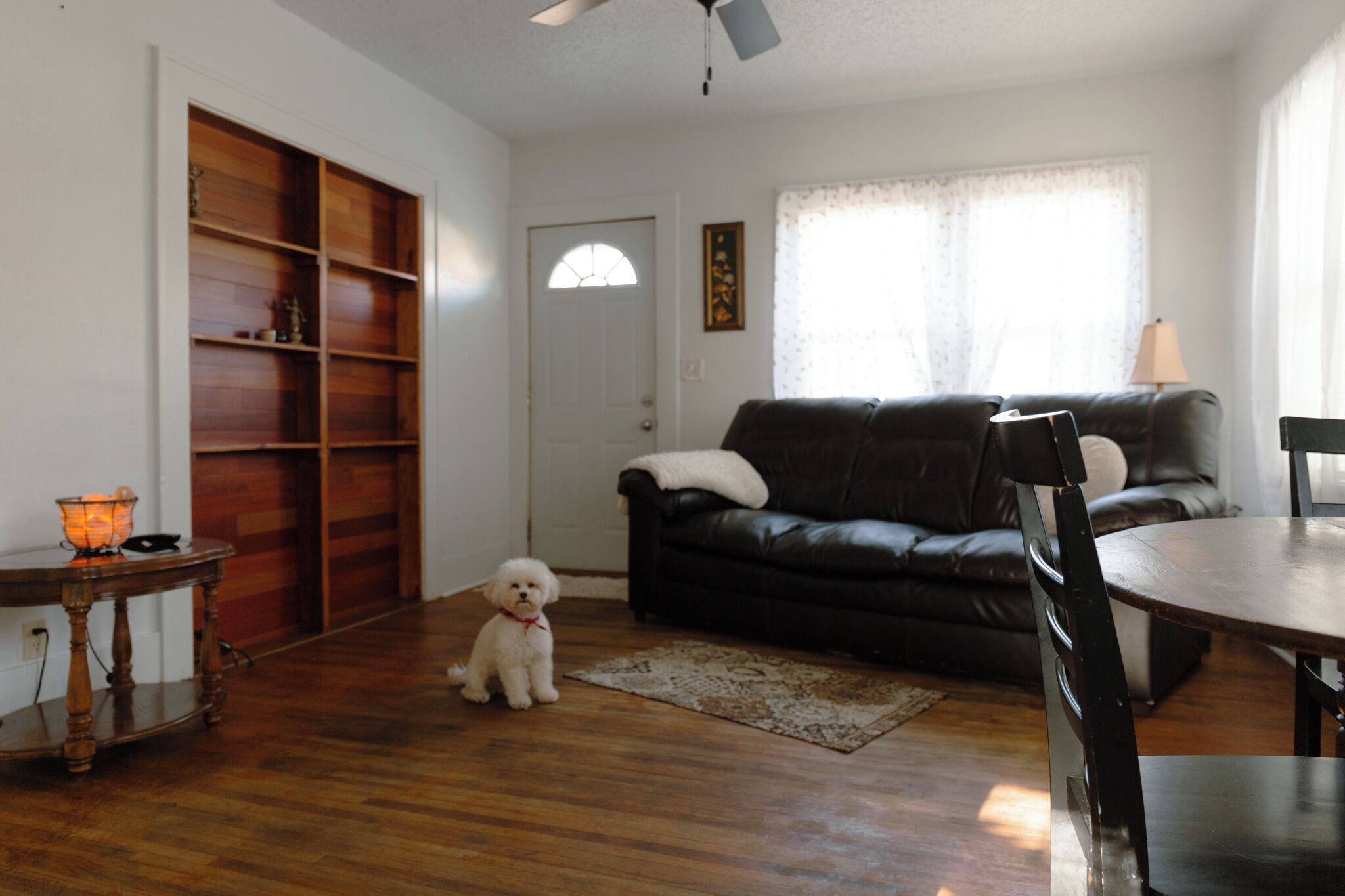A living room with a black leather sofa, a wooden floor, a small table with a candle, and a white dog.