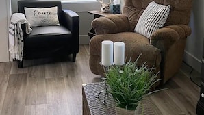 A cozy living room with a brown armchair, a black armchair, a window with blinds, a lamp, and a small table with candles and a plant. - East Side Motel & Cabins (Mobridge)