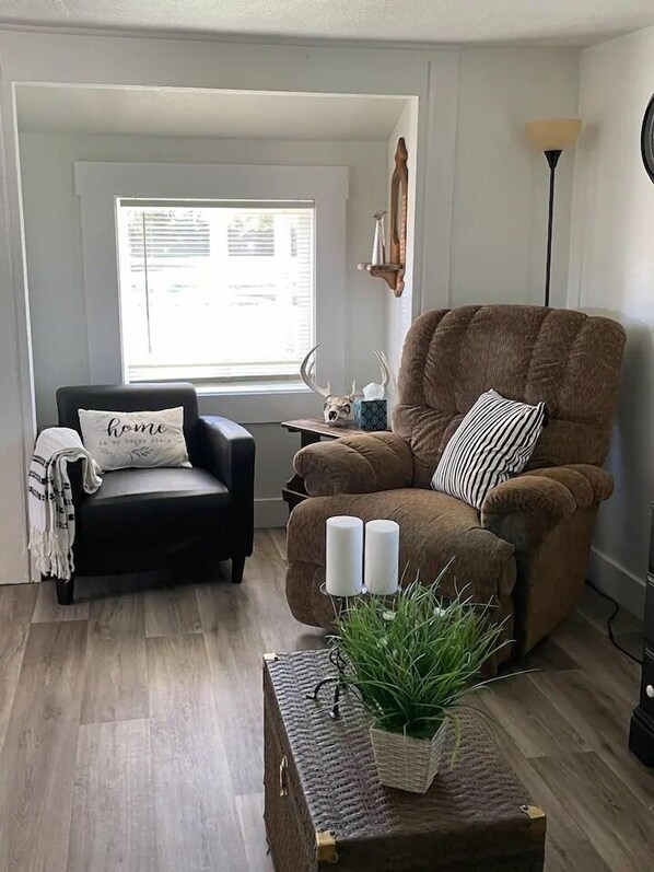 A cozy living room with a brown armchair, a black armchair, a window with blinds, a lamp, and a small table with candles and a plant.