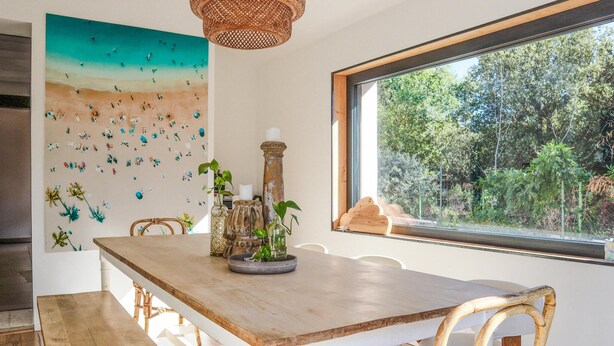 A dining area with a wooden table, wicker chairs, a large painting of a beach scene, and a view of trees through a window. - Beautiful home in Pernes-les-Fontaines (Pernes-les-Fontaines)