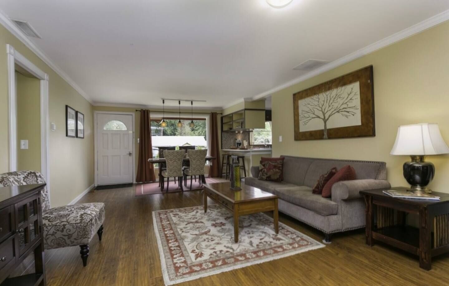 A living room with a sofa, coffee table, and patterned rug. There is a dining area with chairs and a table, and a kitchen in the background.