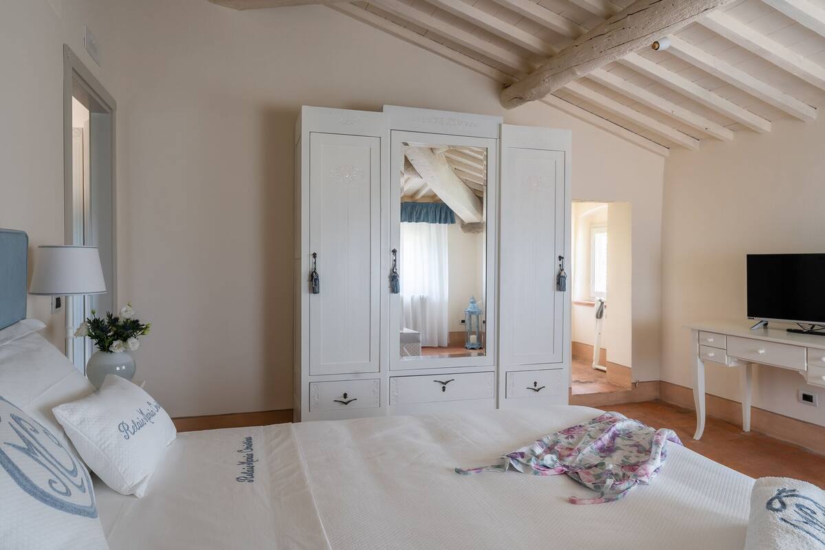 A bedroom with a bed, a white wardrobe, a desk with a TV, and a mirror.