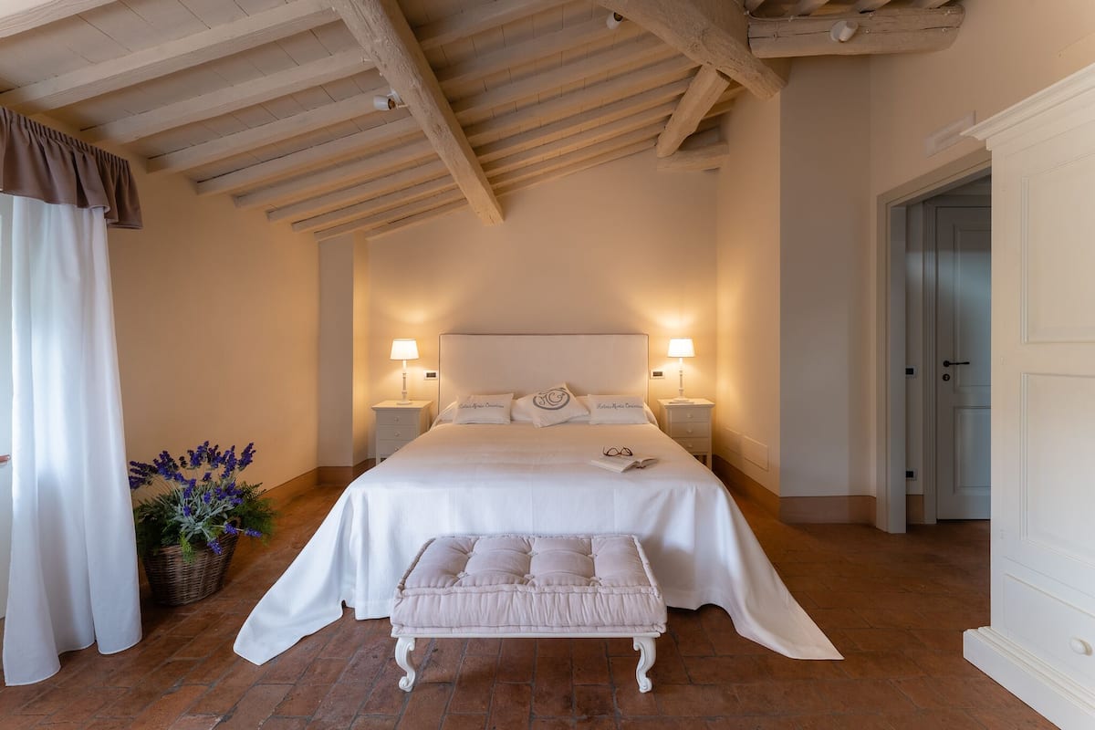 A bedroom with a large bed, two bedside lamps, a white bench, and a potted plant.