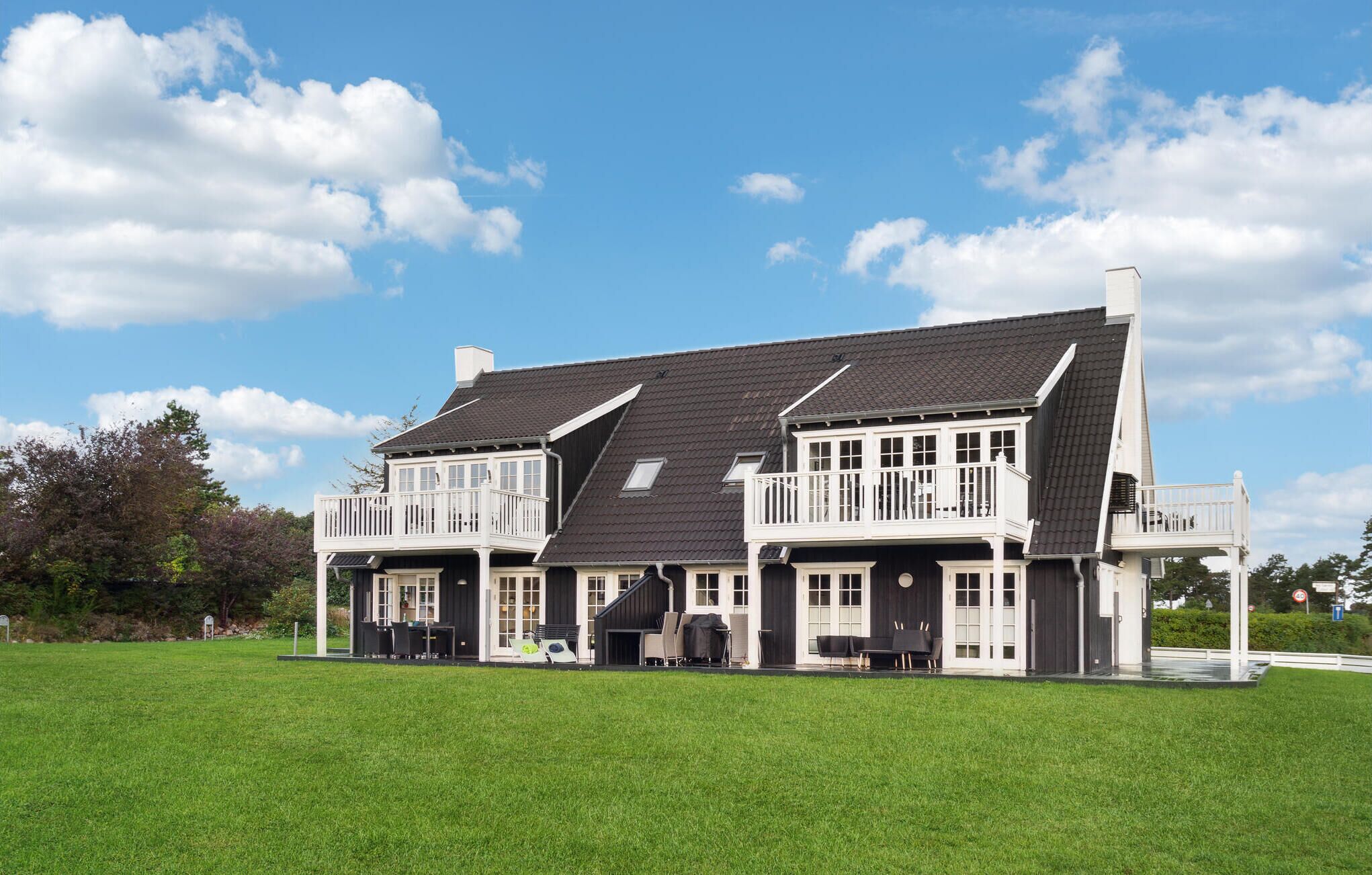 Een groot, twee verdiepingen tellend huis met een donker dak en een witte gevel, met meerdere balkons en een ruime voortuin.
