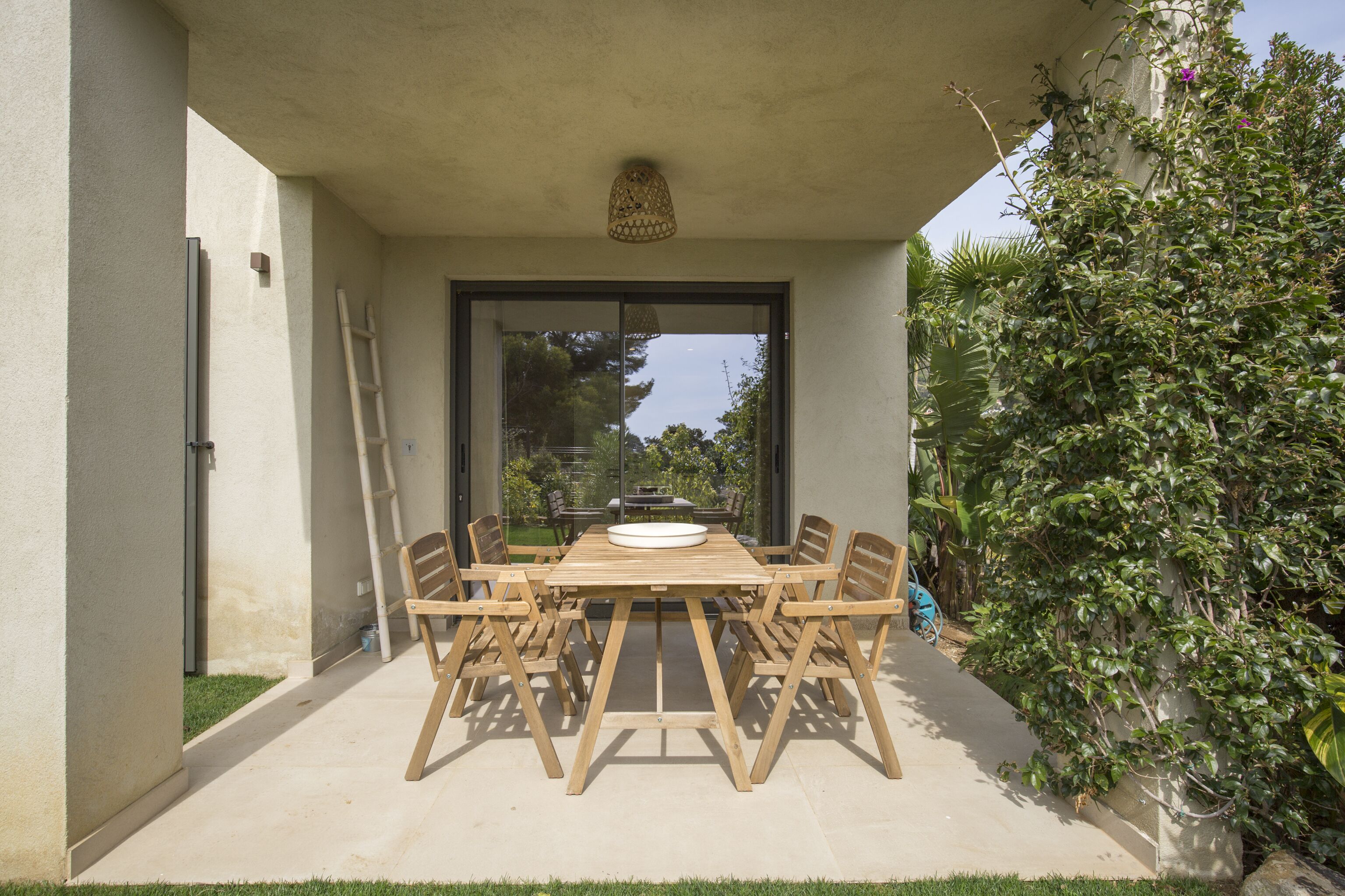 A patio with a wooden table and chairs, surrounded by a concrete floor and a glass door leading to a garden.