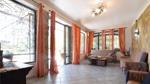 A spacious living room with a sofa, armchairs, and a coffee table. Large windows with curtains allow natural light to fill the room.