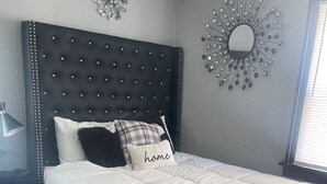 A bedroom with a tufted headboard, a bed with white bedding, a pillow with the word "home," and a wall mirror. - The River Green Getaway
 (Fort Wayne)