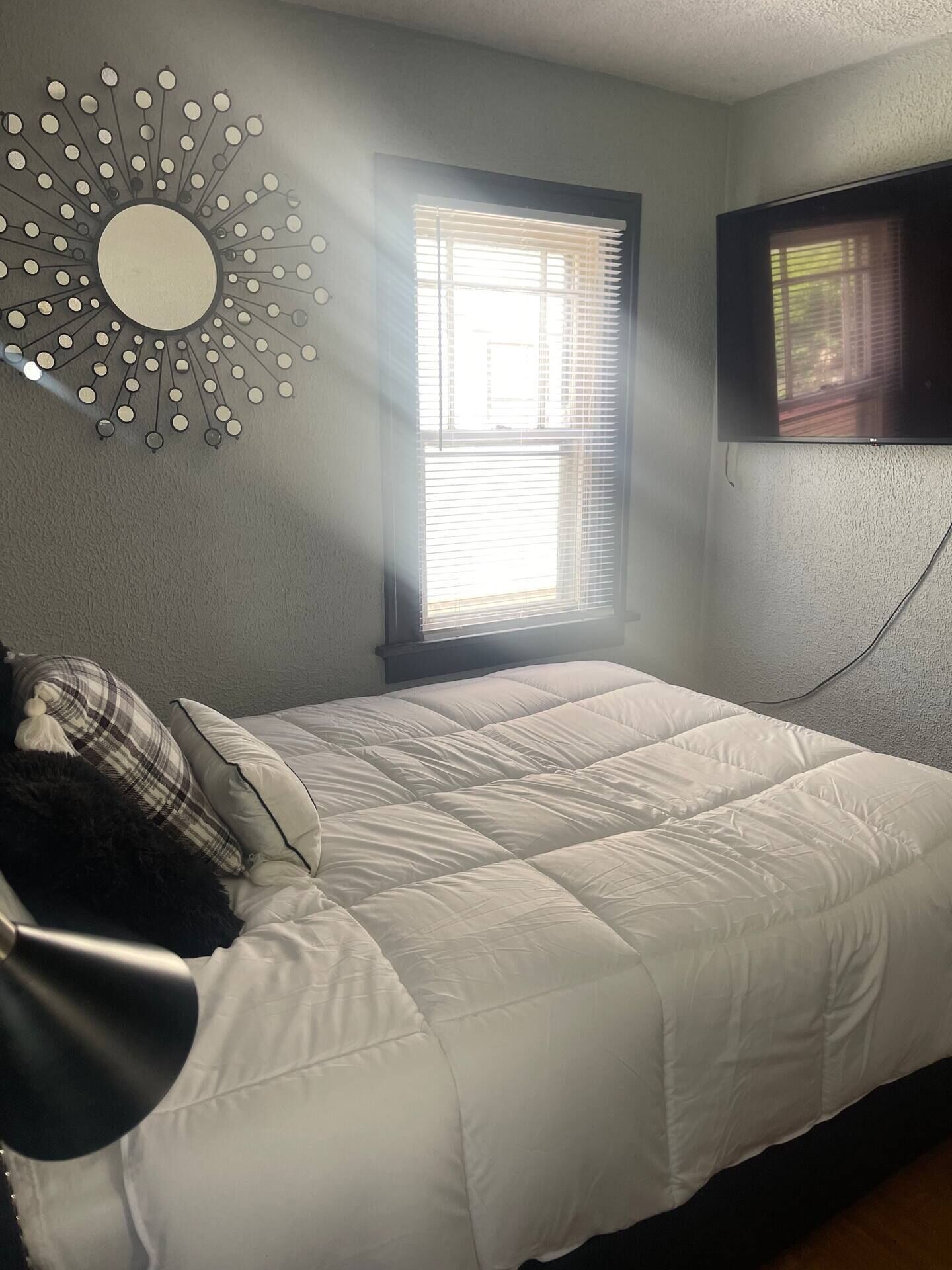 Una camera da letto con un letto, uno specchio fissato al muro, una finestra con persiane e un televisore montato sulla parete.