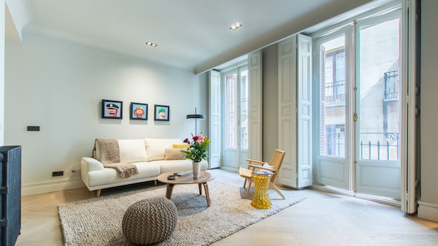 A spacious living room with a white sofa, a coffee table, and a chair. There are large windows with a view of buildings outside.