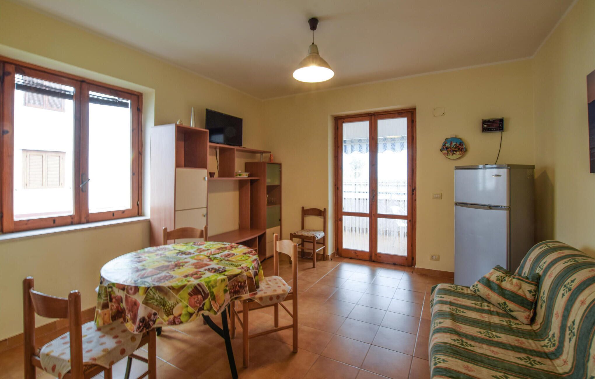 A dining area with a round table covered in a fruit-patterned tablecloth, wooden chairs, a wooden cabinet with shelves, a television on top, and a refrigerator in the corner.