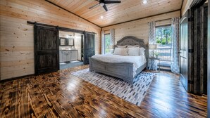 A bedroom with a wooden ceiling, a bed, a ceiling fan, and a view of the outdoors.