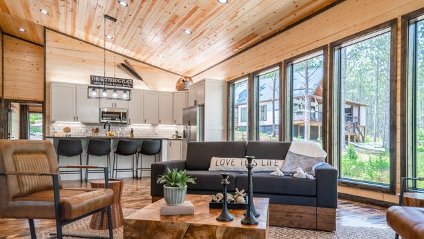 A modern interior with a wooden ceiling, a kitchen area, a living room with a sofa and a coffee table, and large windows offering a view of the outdoors.