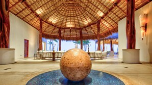 Un'ampia piscina interna con pavimento a mosaico, circondata da tavoli e sedie sotto un tetto di paglia.