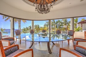 A glass-top dining table surrounded by chairs, with a view of the ocean and palm trees through large windows. - Lower Palmilla: Oceanfront room with pivate pool and jacuzzi (La Paz)