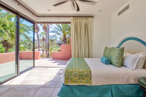 Una habitación de hotel con cama, ventilador de techo, ventana grande y vistas a palmeras.