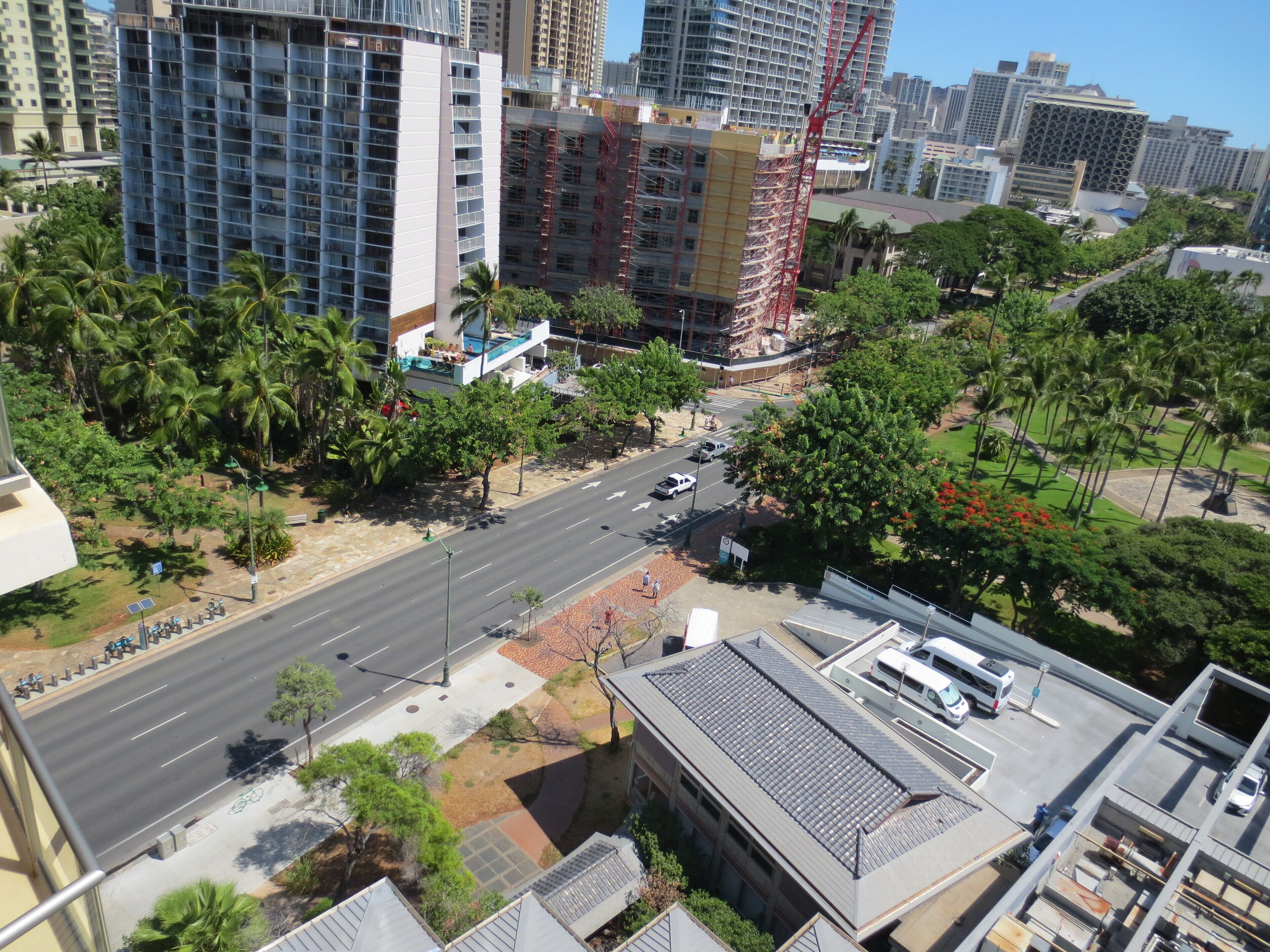 Un edificio alto con varios pisos, una calle con coches y zonas verdes circundantes.