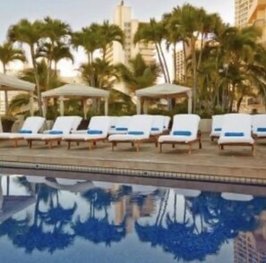 A poolside area with lounge chairs and umbrellas, surrounded by palm trees and buildings. - Amazing view in the beautiful Luana Waikiki! (Honolulu)