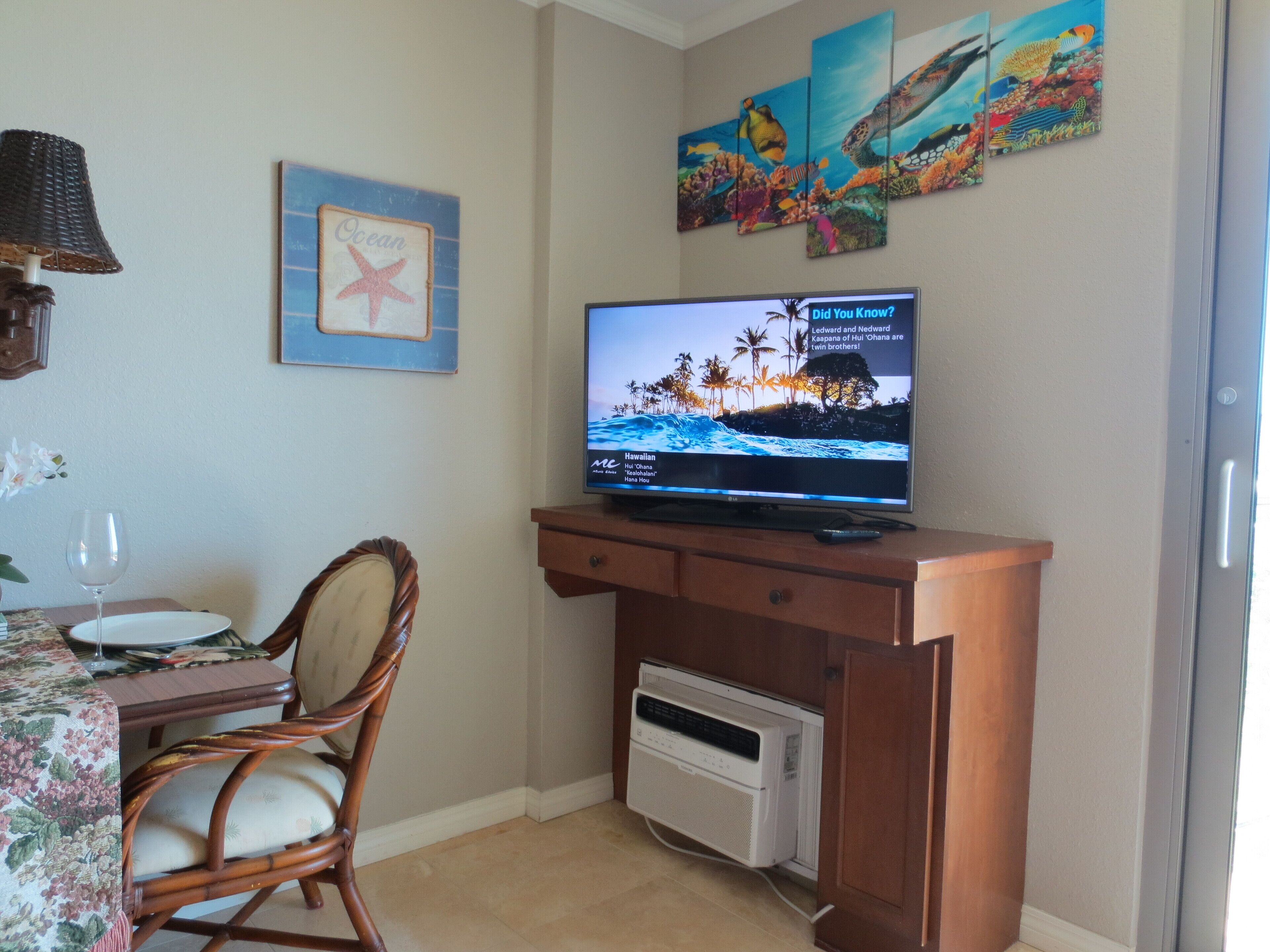 A living room with a flat-screen TV on a wooden stand, a dining table with chairs, a wall-mounted air conditioner, and a framed picture of a starfish.