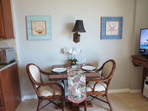 A dining area with a table set for two, featuring a floral tablecloth, two white plates, two wine glasses, and a vase with white flowers. There are two wicker chairs, a wall-mounted lamp, and two framed pictures on the wall. - Amazing view in the beautiful Luana Waikiki! (Honolulu)