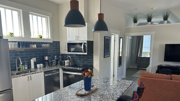 A modern kitchen with granite countertops, white cabinets, and a central island. There is a dining area with a brown sofa and chairs.