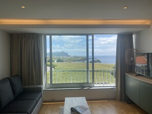 Una sala de estar moderna con un amplio ventanal que ofrece vistas a un paisaje costero, un sofá negro, una mesa de centro y un televisor sobre un mueble.
