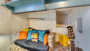 Quarto rústico com cama, almofadas, um violão e uma parede de pedra.