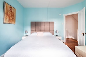 Un dormitorio con una cama grande, dos mesitas de noche con lámparas, un cabecero y una puerta que da a otra habitación.
