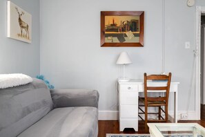 A room with a grey sofa, a white desk with a lamp, a wooden chair, and two paintings on the wall.