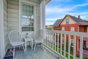 A porch with two white chairs and a small table, a white railing, and a view of a red brick house across the street.