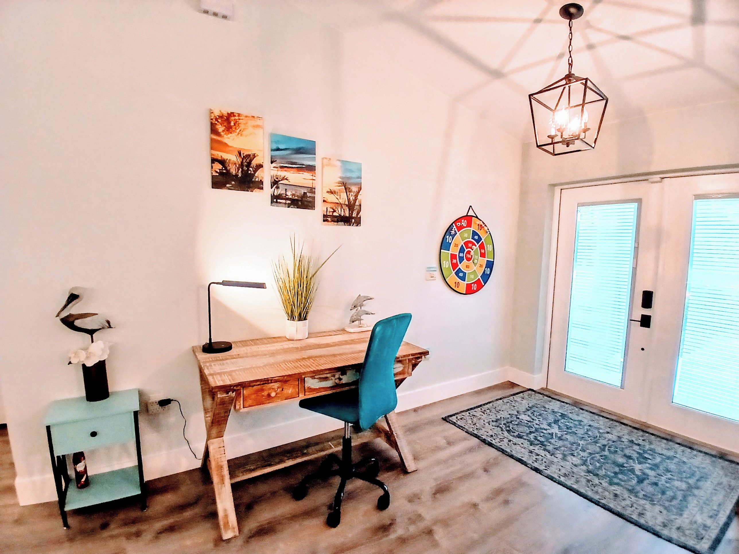 Une pièce avec un bureau en bois, une chaise de bureau bleue, une lampe, une plante en pot, des œuvres d’art murales, un jeu de fléchettes et une porte avec des stores.