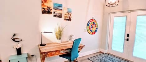 A room with a wooden desk, a blue office chair, a lamp, a potted plant, wall art, a dartboard, and a door with blinds.