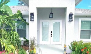 A white house with a front door, two lantern-style lights, and a small garden with various plants and flowers.